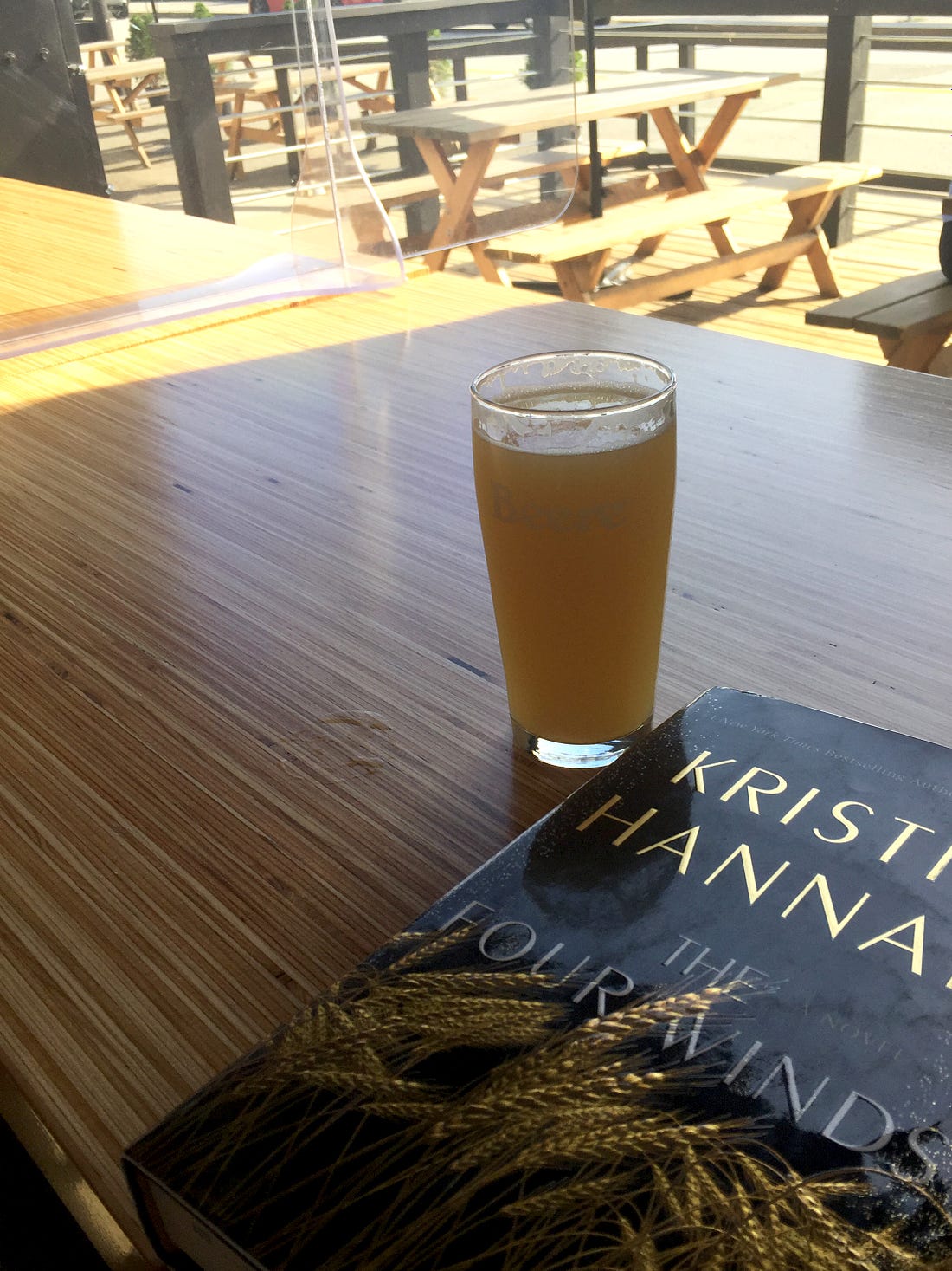 a wooden table with a view of a patio with picnic tables, and the street beyond. On the table is a glass of hazy IPA and a black hardcover book, Kristin Hannah's "The Four Winds".