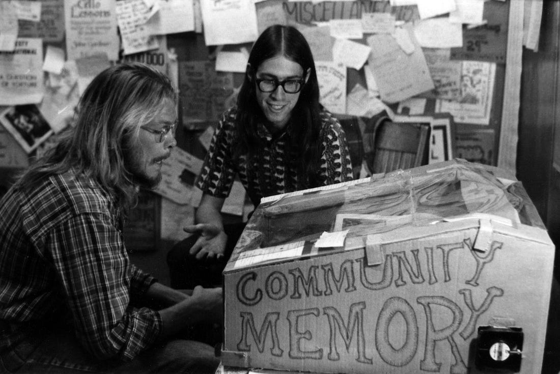 A black and white photograph of two white men with long hair and patterned shirts sitting in front of a physical bulletin board. One of the men is using a computer terminal within a cardboard case with “COMMUNITY MEMORY” written on the side