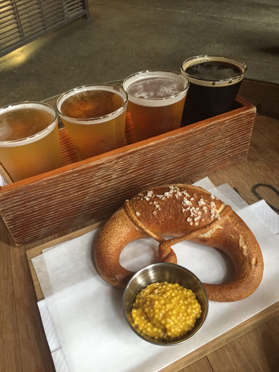 a flight of four beers in a wooden box, with the lightest at the left and a dark stout at the right. In front of them is a twisted soft pretzel with big salt flakes, and a ramekin full of grainy mustard.