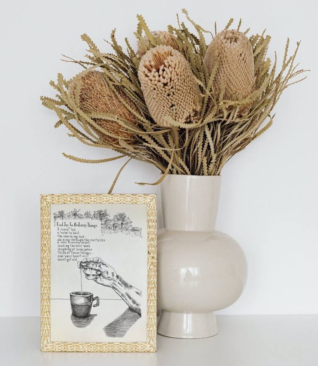 A photo of the mock-up of the artwork, Find Joy In Ordinary Things Illustrated Poem in a muted gold, weaved frame, beside it is a light beige coloured vase with dried flowers in autumn colours of golden yellow, light brown, and cream.