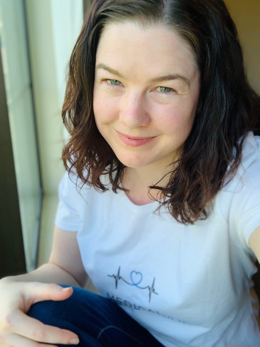 Shasta Kearns Moore, a brown-haired, blue-eyed white woman smiles at the camera wearing a white T-shirt with the Medical Motherhood logo and blue jeans.