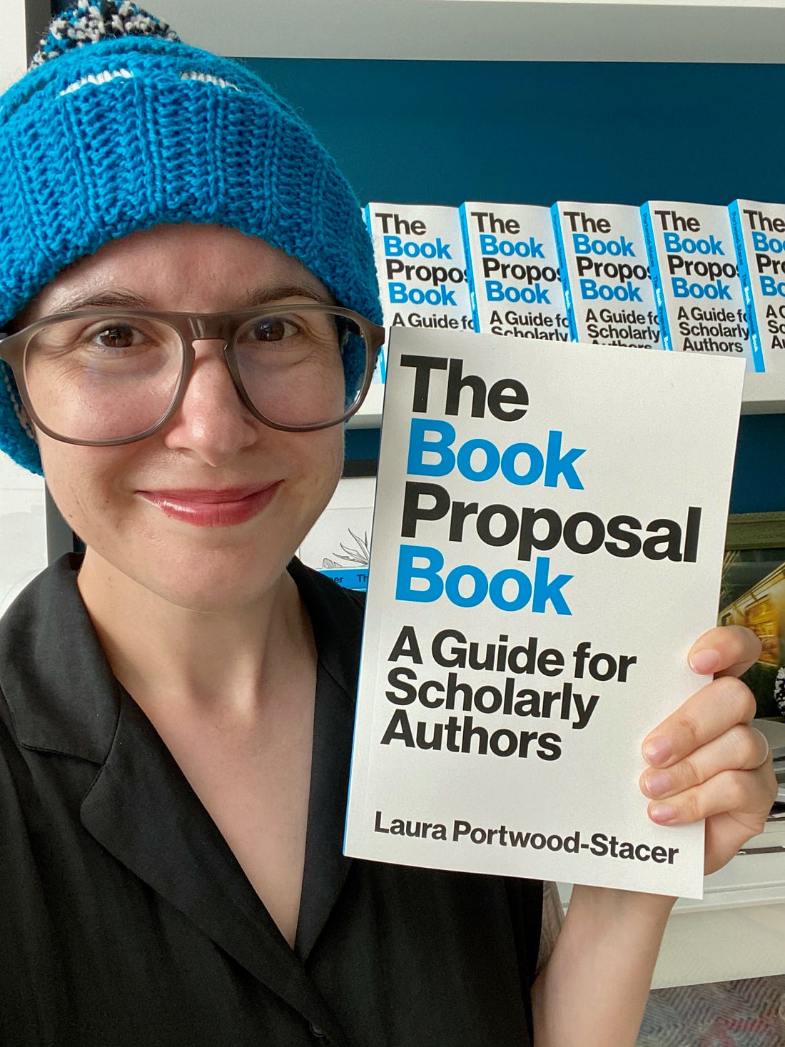 A photo of me holding up The Book Proposal Book and wearing a knitted cap in colors that match the book's cover (bright blue, white, and black)
