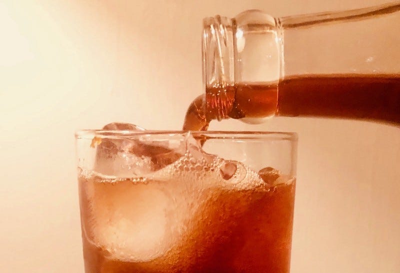 Closeup of a reddish-brownish liquid being poured from a clear bottle into an almost-full glass with ice in it.