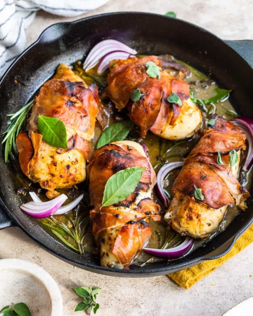 prosciutto wrapped chicken in a cast iron skillet on top of a striped dish cloth