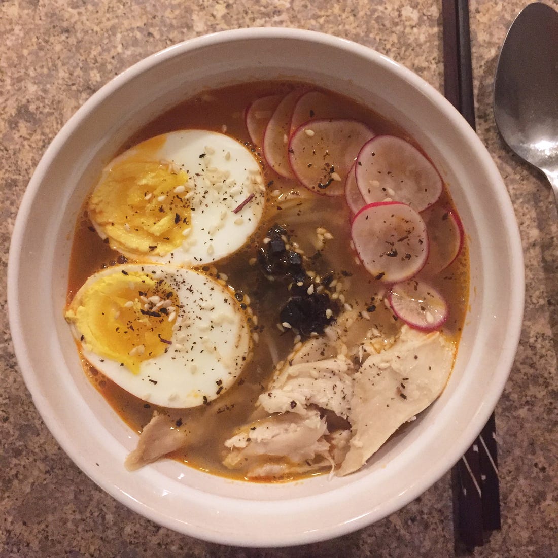 A large white bowl full of ramen. The noodles are just visible under a boiled egg, a pile of chopped black garlic, thin radish slices, and shreds of chicken. Dried shiso and toasted sesame seeds are sprinkled over the top, and to the right of the bowl are a large spoon and a set of dark wooden chopsticks.