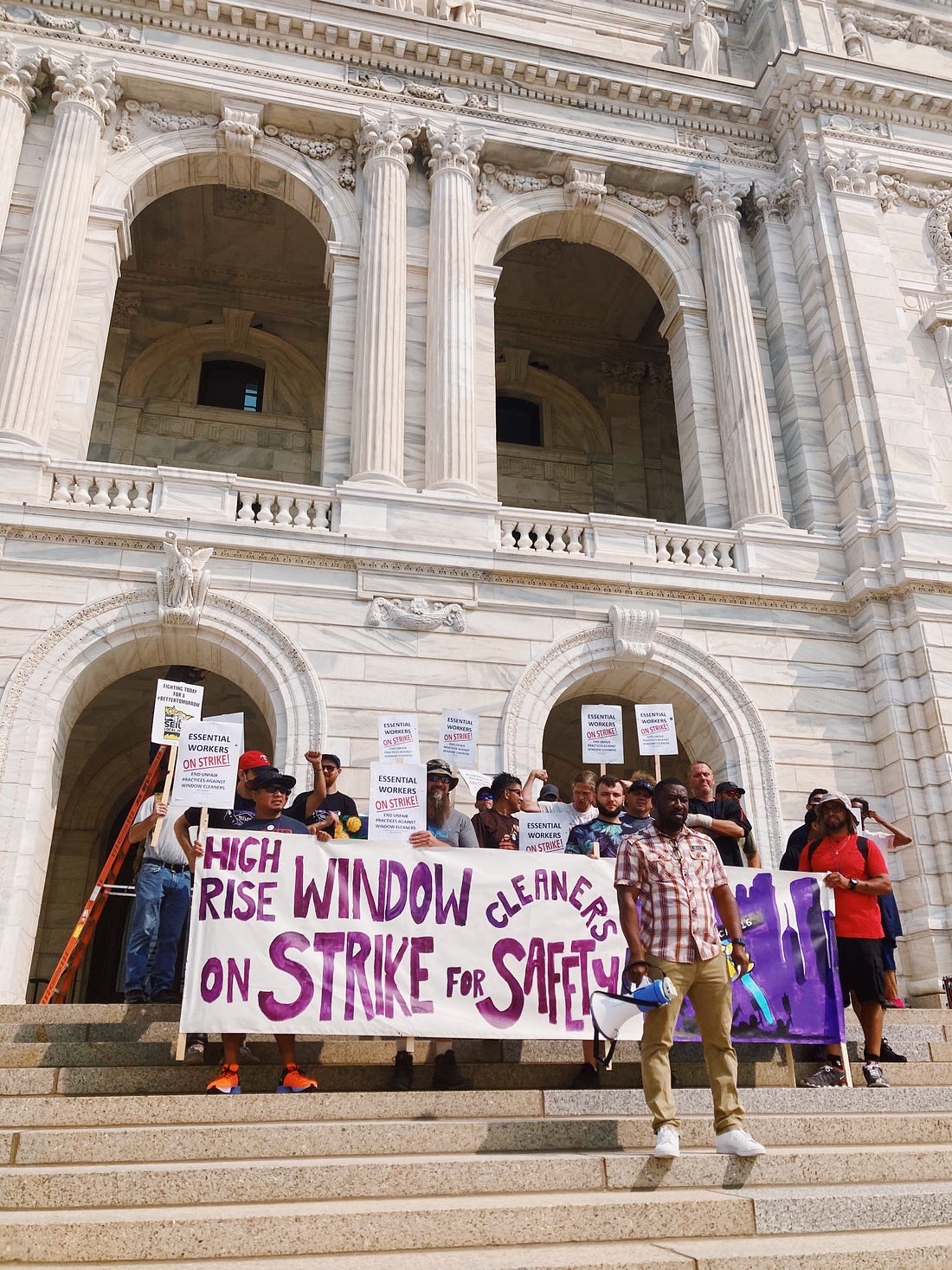 a group of people holding picket signs and a banner reading "high rise window cleaners on strike for safety" in purple letters stand in front of the white marble capitol building steps