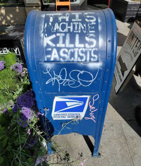 Photo of a blue mail box that has words stenciled in white that say, "this machine kills fascists"