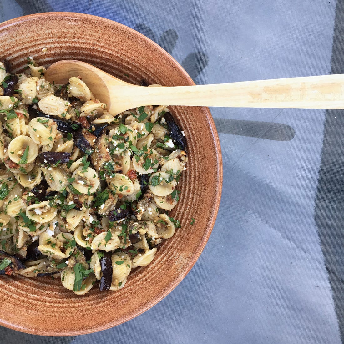 a textured brown ceramic serving bowl filled with orecchiette pasta salad with charred eggplant slices, parsley, sun-dried tomato, and feta visible.