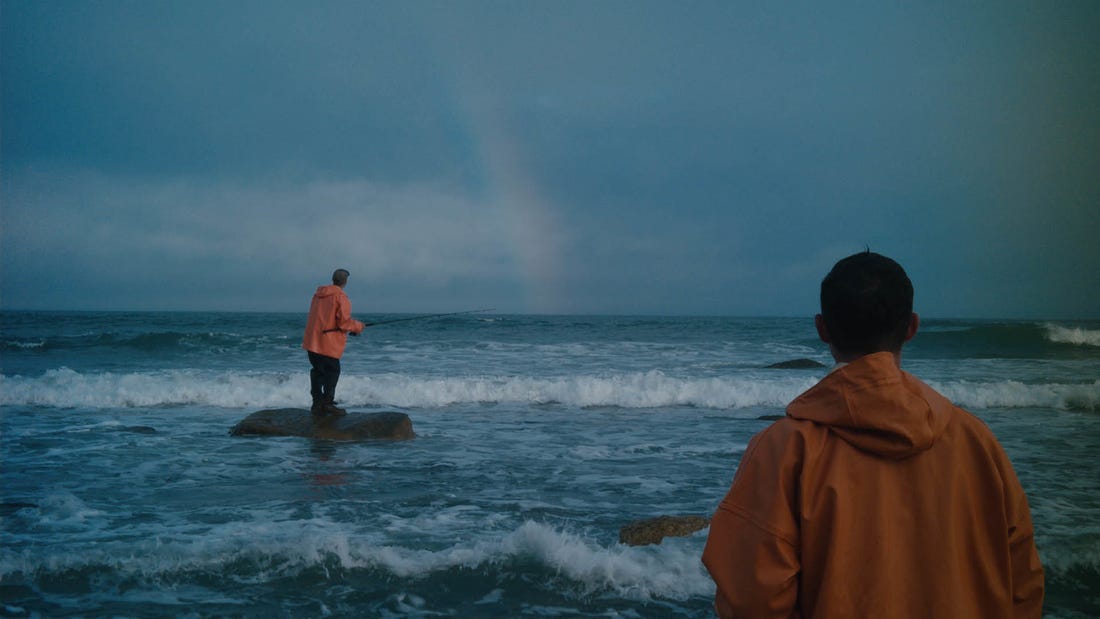 An official still from the film 'Somewhere with No Bridges', where two fishermen stand off a beach, fishing into the ocean during the day, with a rainbow in front of them in the distance.