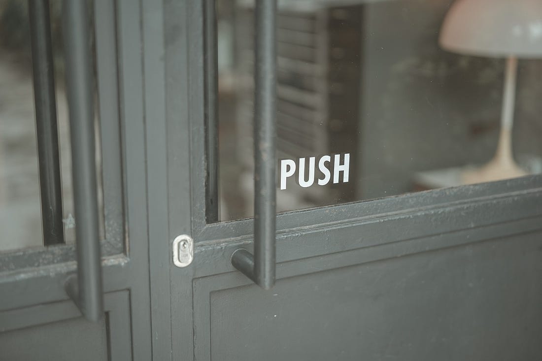 The word PUSH shows in white letters on the glass window of a heavy gray door with a small silver keyhole and a while lamp inside the door.