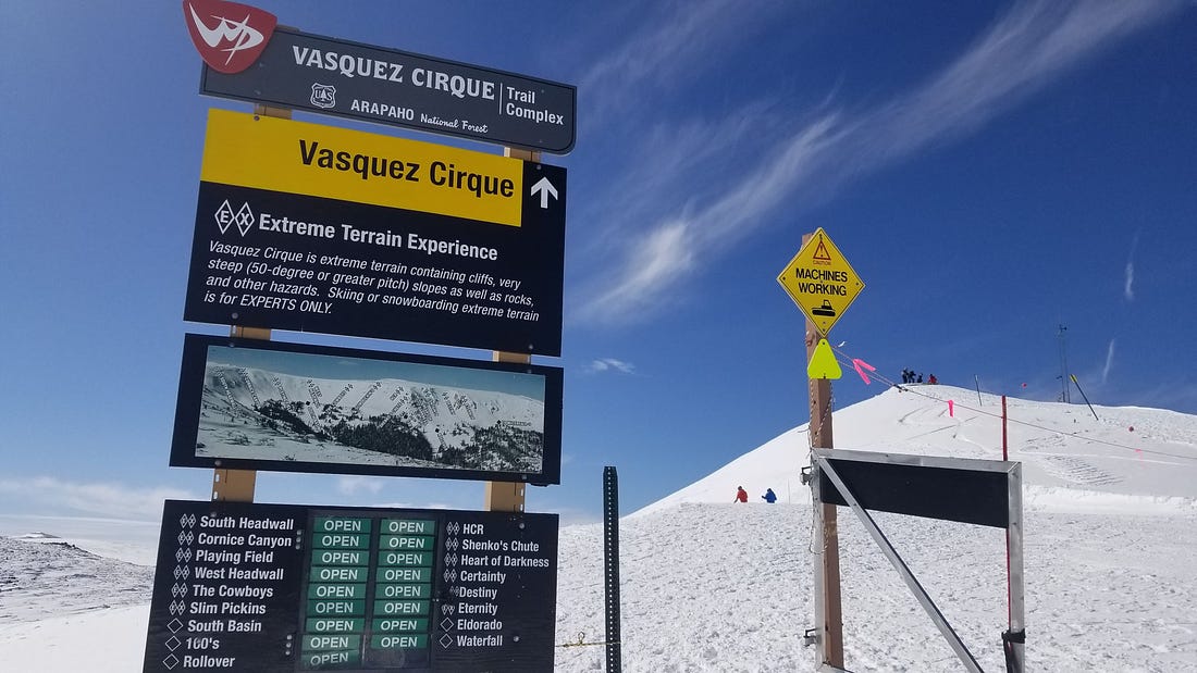 A sign stands next to an open gate atop the mountain. Above, the sky is a hazy blue. Pristine snow, packed in with footprints coats the mountaintop.