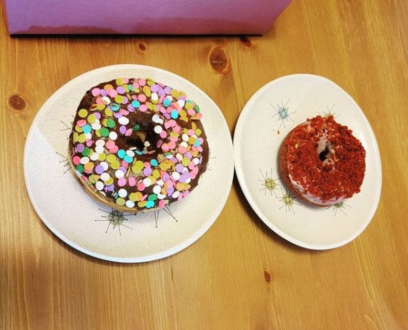 photo of two donuts, each on a small vintage dessert plate. One donut has chocolate frosting and round confetti rainbow sprinkles, the other is a red velvet donut with a red crumbly topping