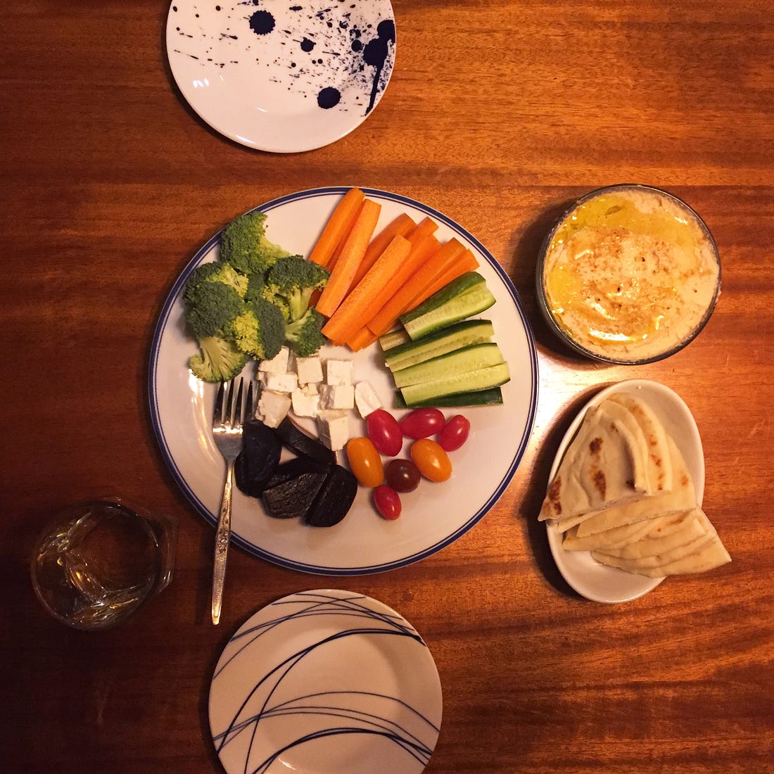 A large plate of broccoli, carrot and cucumber sticks, and grape tomatoes with cubes of feta and pickled beets. To its right is a small dish of flatbread wedges, and a glass container of white bean hummus dusted with paprika and coated in olive oil. Two small plates sit on either side of the food.