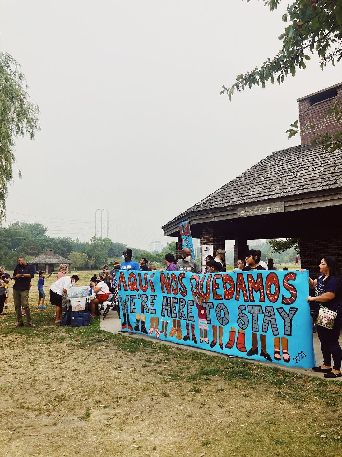 a group of people stand in front of a shelter at a park holding a bright blue sign with red and grey letters reading "aquí nos quedamos/we're here to stay" above paintings of feet