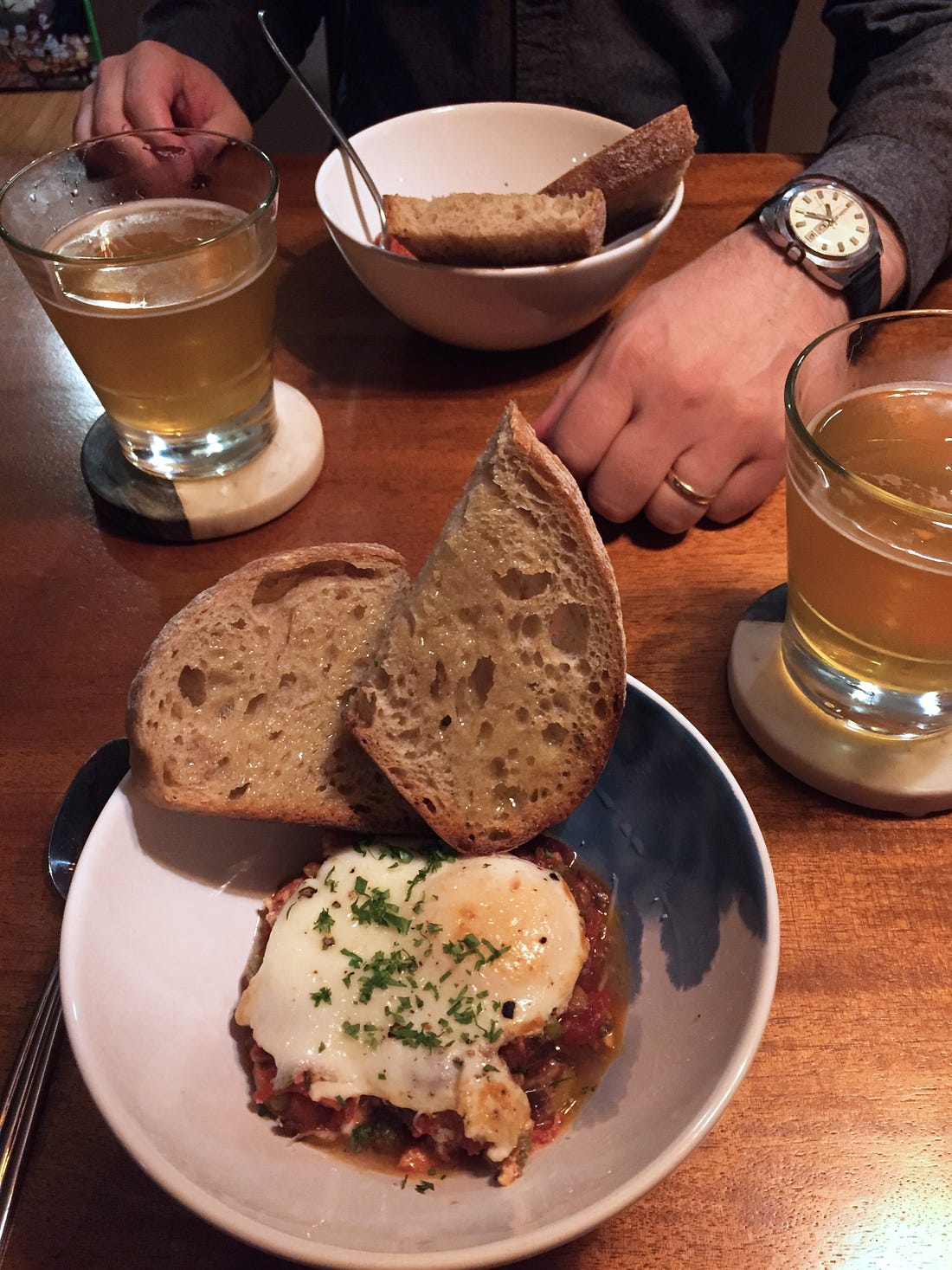 A table setting with two bowls of puttanesca, each with two slices of toast at the side and a poached egg and a sprinkle of parsley on top. Two glasses of beer sit on coasters to the sides of the bowls, and Jeff's hands are visible at the other side of the table.