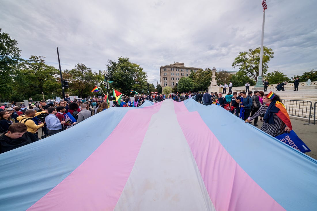A large trans flag is unfurled on the street in front of the Supreme Court, by dozens of protestors
