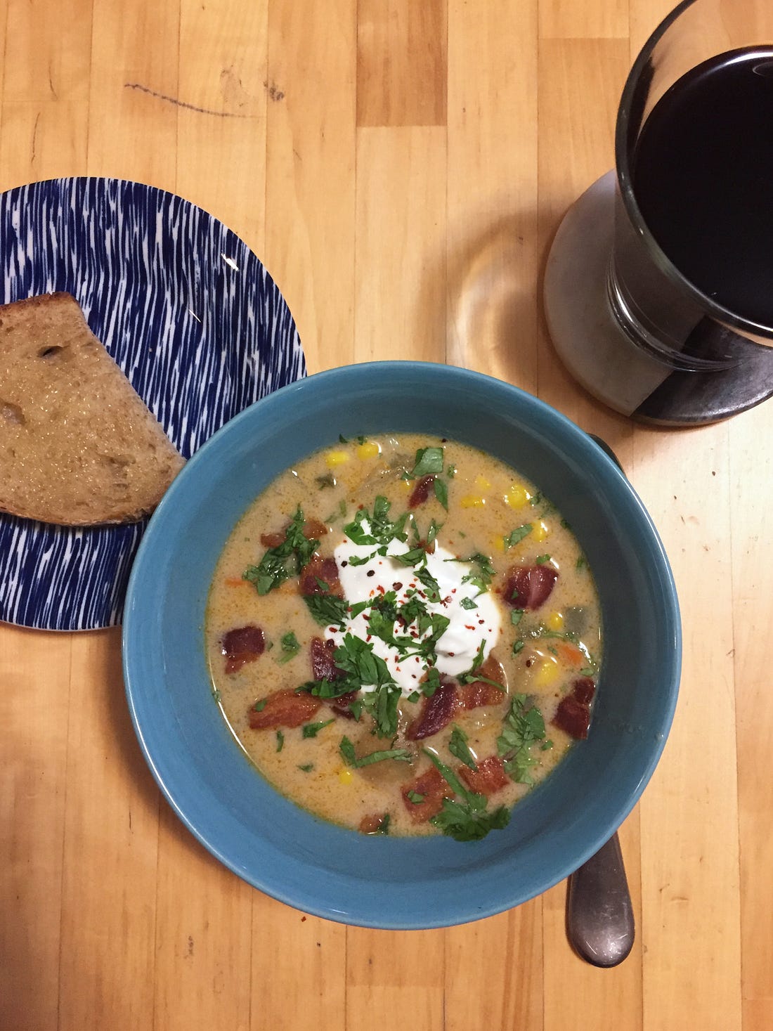 a blue bowl full of corn chowder, and topped with pieces of bacon, cilantro, and a dollop of crema. To the left, a piece of toast on a blue plate. To the upper right, a glass of dark beer.