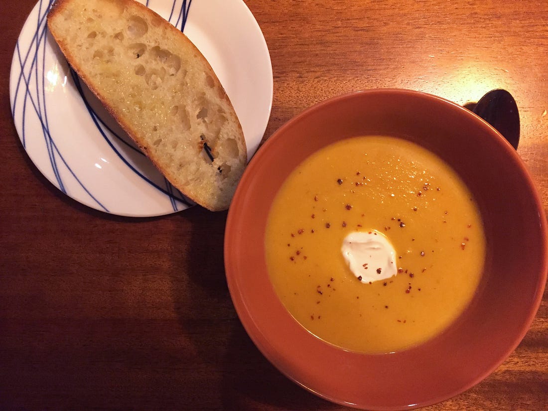 An orange bowl full of dark yellow squash soup, with chili flakes dusted overtop, and a dollop of sour cream in the centre. Beside it to the upper left is a small plate with a slice of buttered sourdough.