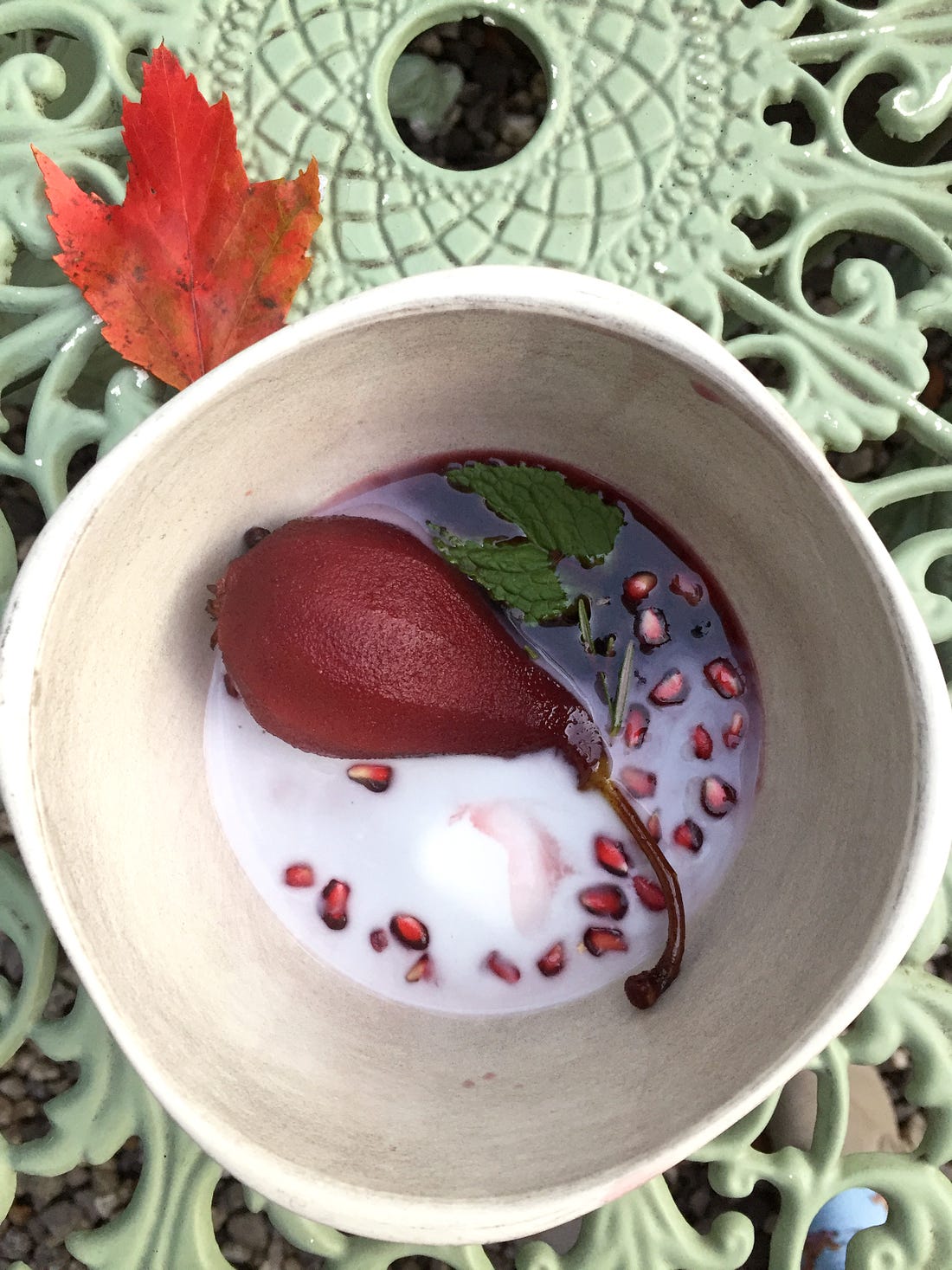 On an outdoor table, a small white bowl sits with a maple leaf at its upper corner. In the bowl is a dark red poached pear sitting in wine an coconut cream with pomegranate seeds flecked throughout, and mint and rosemary at the edge of the bowl.