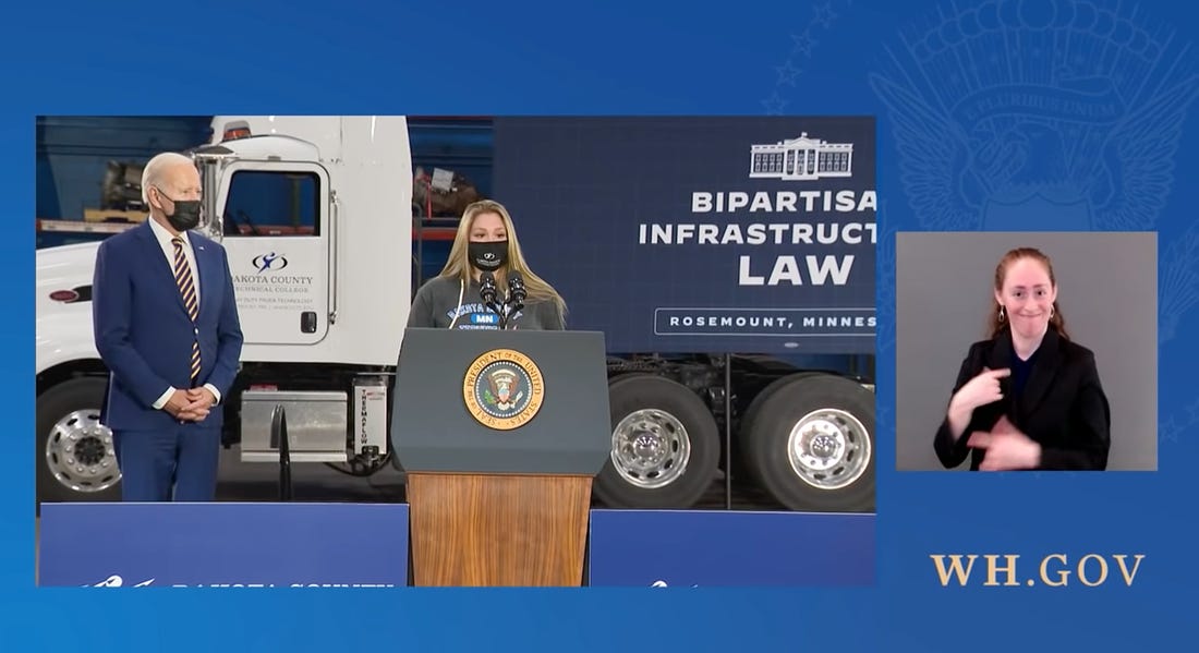 screenshot of presidential livestream, blue background with one screen showing a blonde woman with black mask standing behind a podium, president biden wearing a blue suit and black mask standing beside her, in front of a semi truck with a poster reading bipartisan infrastructure law, another screen shows an ASL interpreter