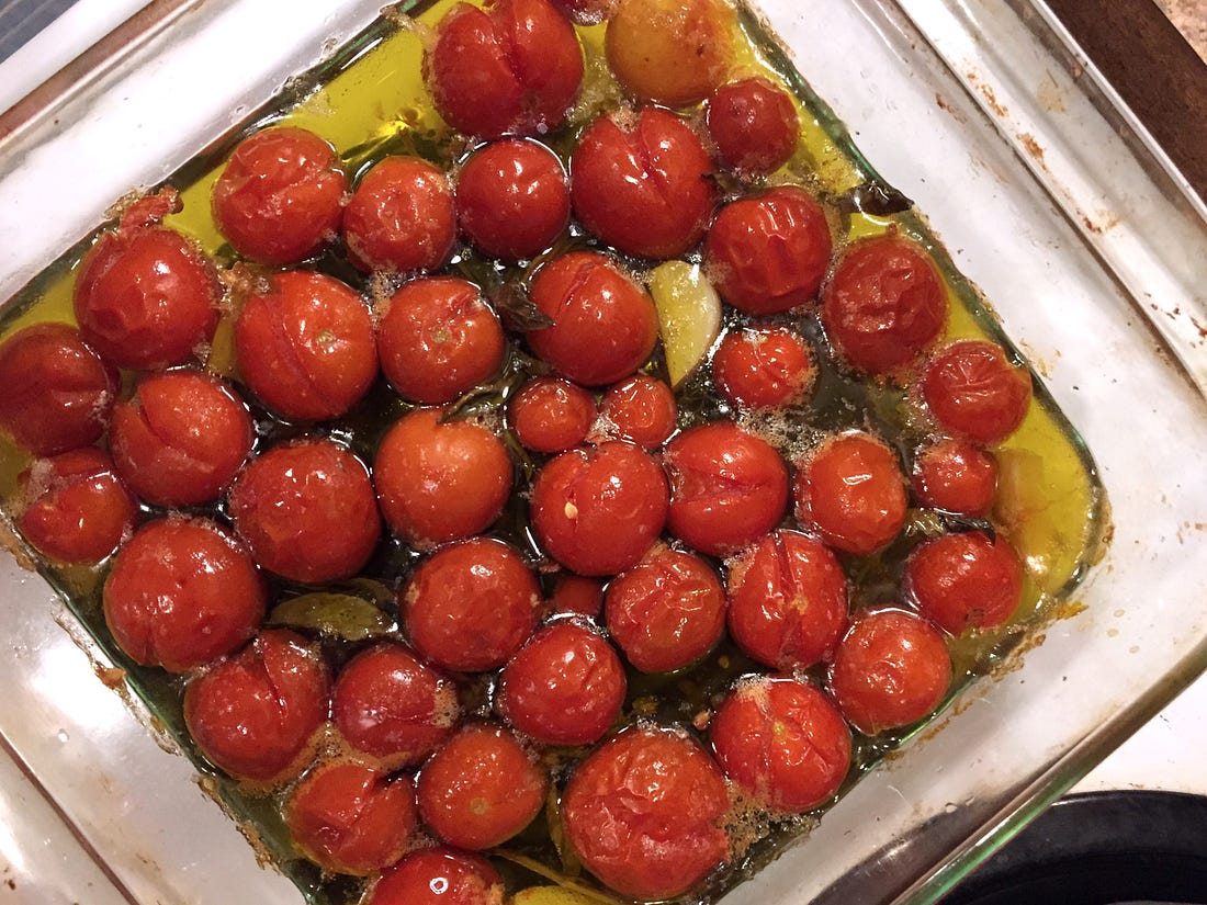 from above, a clear square baking dish full of lightly bubbling olive oil, cherry tomatoes that have split in places, and a few garlic cloves and basil leaves.