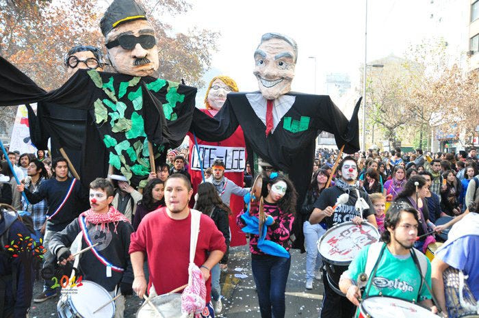 A slightly overexposed, washed-out digital image of a student protest marching down a wide street, probably in downtown Santiago. In the foreground, young people walk playing large drums and cymbals, a sort of popular marching band known as a "batucada." Just behind them loom four giant papier mache puppets—each two people tall—carried by other students on poles. The two in front are former dictator Augusto Pinochet on the left, and president Sebastián Piñera on the right. They are dressed in all black and grin evilly, pockets full of money. Behind them are two other puppets, partially obscured, and students marching out as far as can be seen.