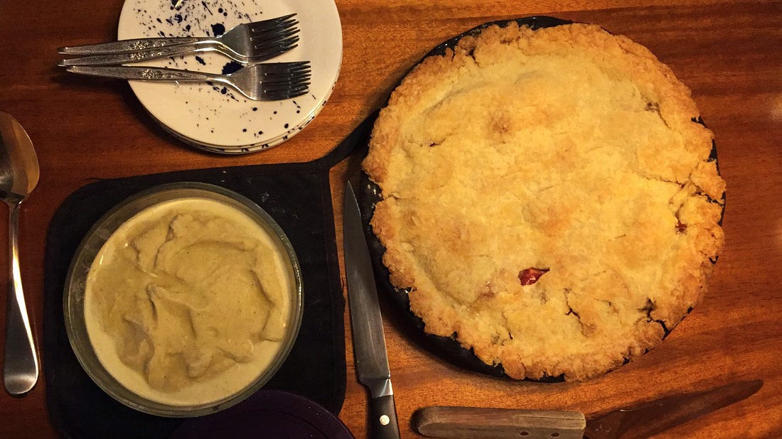 An apple pie in a black pie pan next to a glass container of pistachio ice cream. A stack of dessert plates and forks is visible in the upper corner of the photo, and a pie server in the bottom corner.