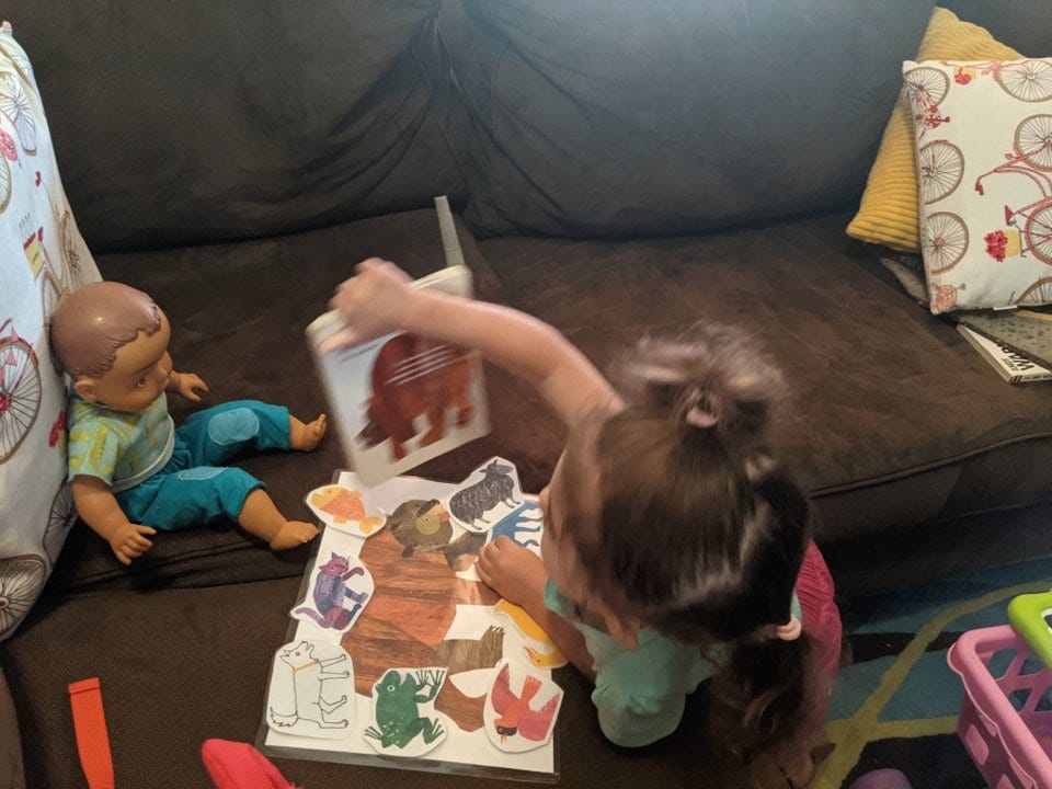 Image of Lila holding a board book up to a babydoll who is sitting on the couch.  A laminated set of animal photos on a paper is in front of her.