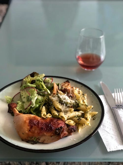 A dinner plate with roast chicken, salad, and pasta nest to silverware and a glass of red wine.