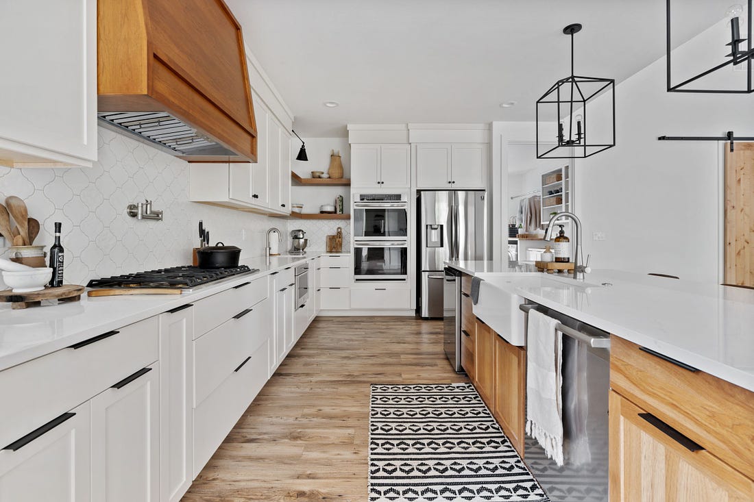 The kitchen galley shows a double oven and fridge at one end. The stainless steel appliances gleam in the white kitchen with black fixtures and wood accents. 