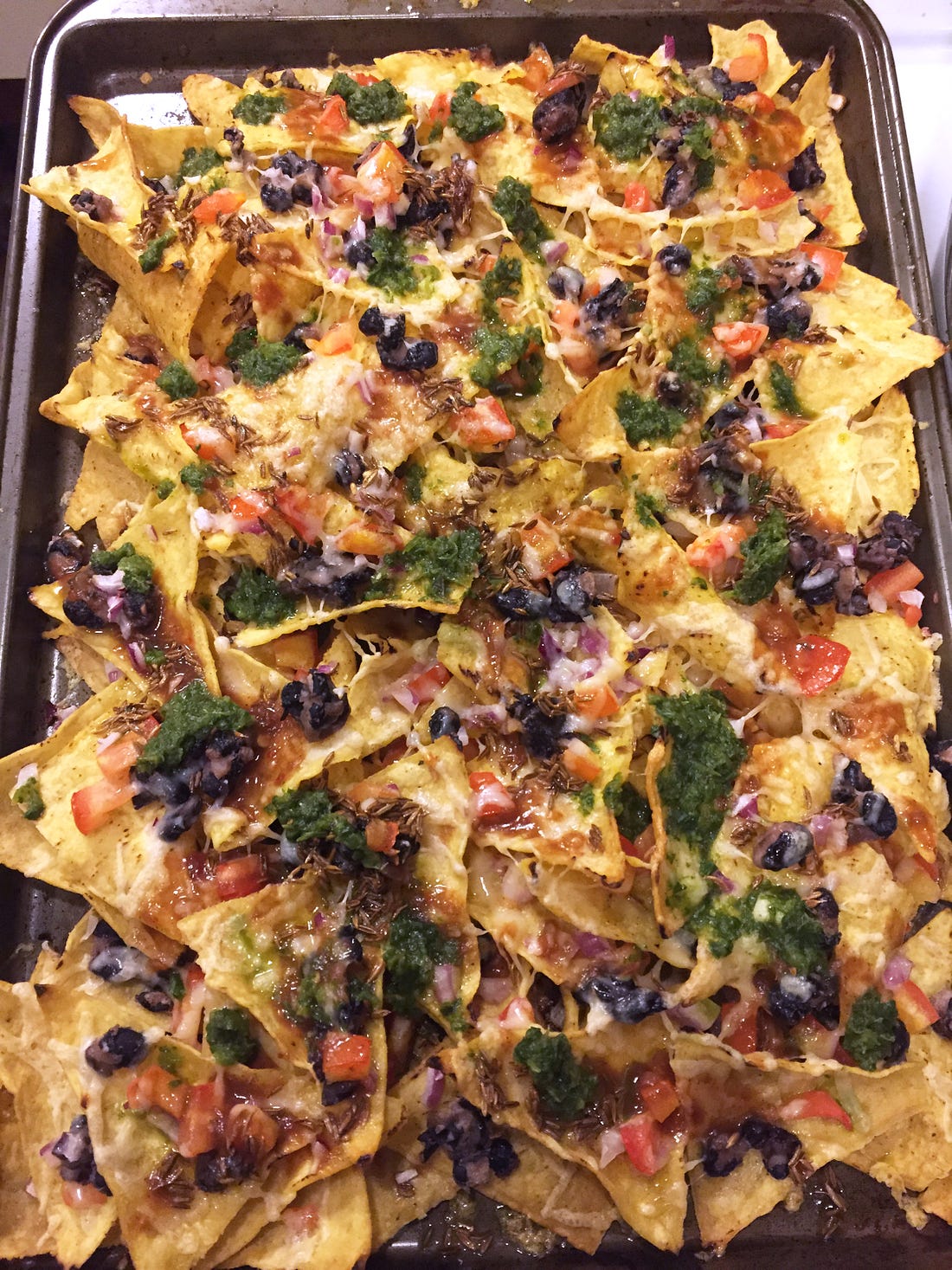 A pan of nachos with tomatoes, black beans, and red onions visible throughout. Bright green drops of the cilantro chutney spatter the top, and patches of cumin seeds can be seen amid the toppings.