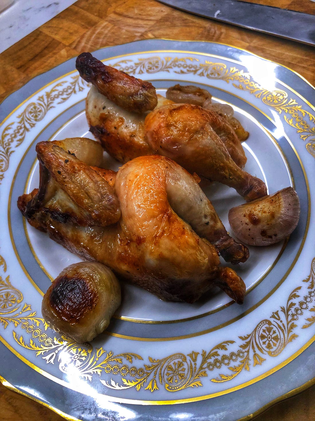 A fancy dinner plate holding two halves of a browned, roasted Cornish hen and roasted shallots.