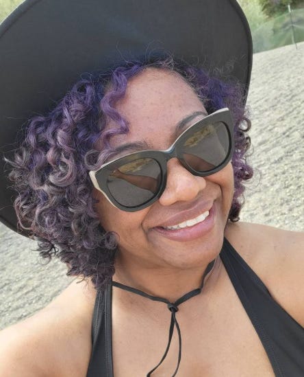 a selfie of Patricia. She is wearing sunglasses, a black sun hat, and a black bikini top. She is smiling.