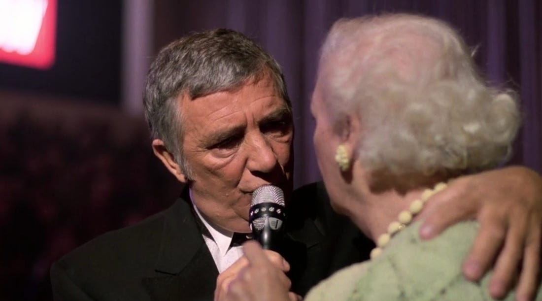 Richard Dawson clutches an elderly woman in a green sweater by the back of her neck and speaks to her up close, holding a microphone. 
