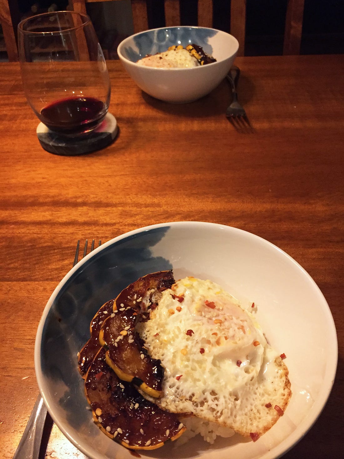 On a wood table, two white and blue bowls sit across from each other, with a stemless glass of red wine at the side. The bowls are filled with rice and topped with half-moons of the hoisin squash, and a fried egg with crispy edges. Sesame seeds and chili flakes are scattered across the top.