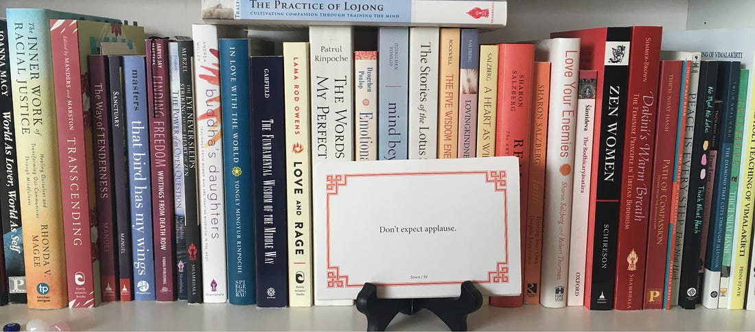 Photo of the lojong slogan card "Don't expect applause" sitting on a white bookcase surrounded by Buddhist books. 