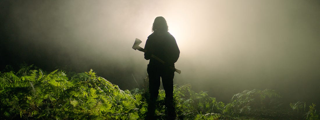 From the film "In the Earth": The silhouette of a man carrying an axe appears through light and fog in the middle of deep forest shrubbery.