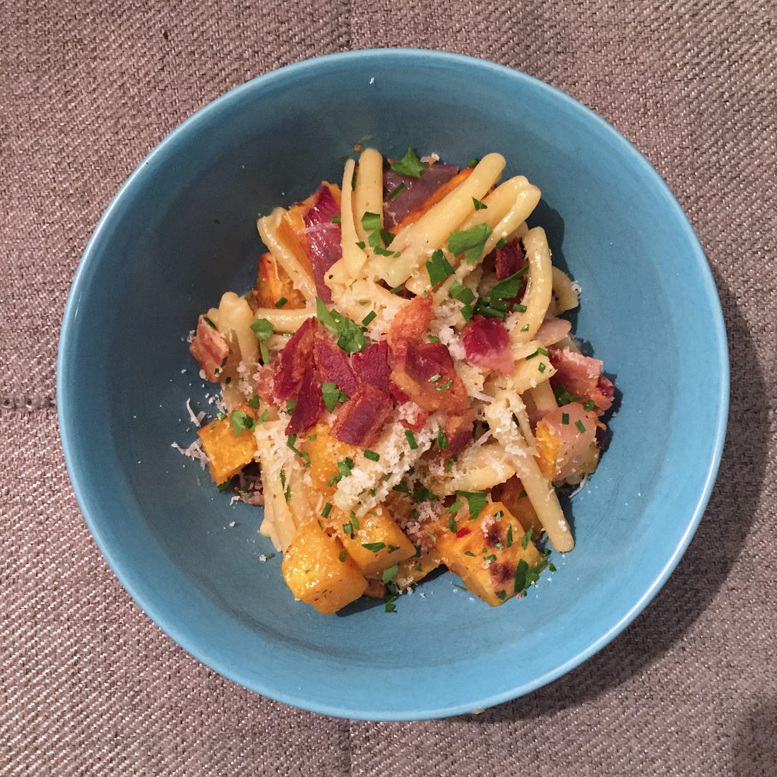 a blue bowl full of casarecce, browned cubes of butternut squash, and pieces of bacon. On top is a dusting of chives and parsley, and parmesan.