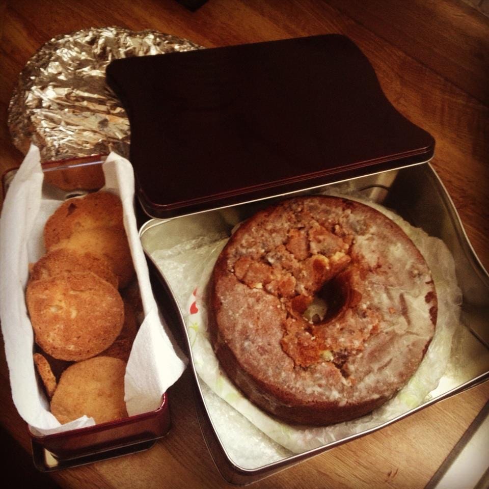 A large brown tin is holding a round cake with melted sugar frosting.  The lid sits off the side of the box.  Another rectangular tin holds several golden brown tea cake cookies