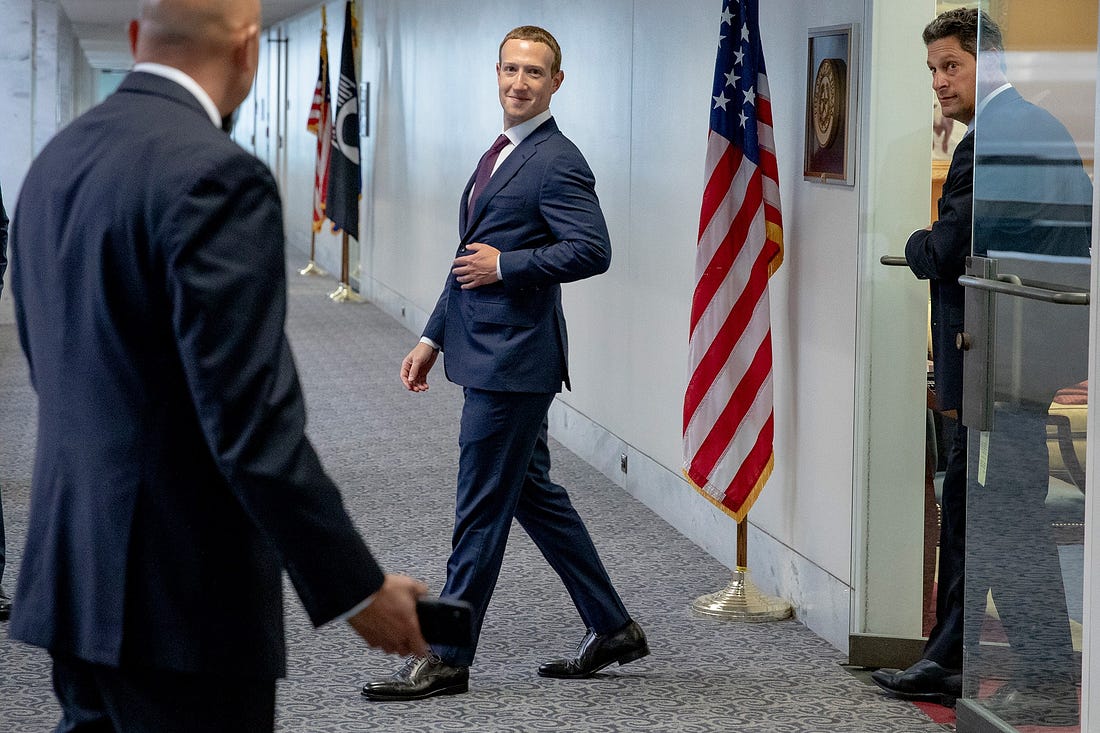 Mark Zuckerberg walks out of a room into a government office hallway next to an American flag.