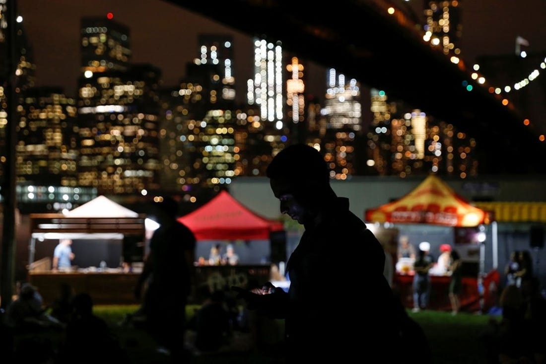 Under a bridge in New York City, cover of nightfall, a man looks at his phone against a background of kiosks and tents offering food and wares after hours.