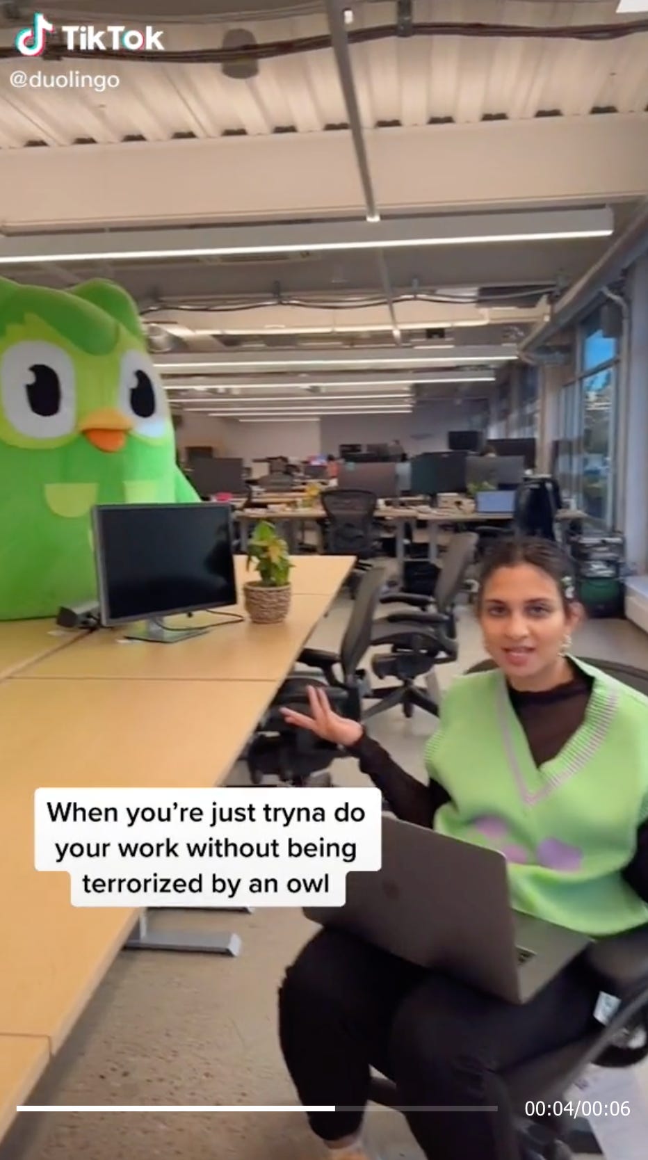 Screenshot of a TikTok video with a woman sitting in the foreground and a green owl costume in the background. Text on screen says "When you're just tryna do your work without being terrorized by an owl."
