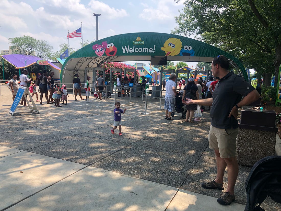 Photo of entrance of Sesame Place and Lila holding her hands out signing for Mark to join her.