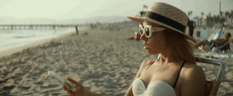 On the beach, Ingrid lowers her sunglasses and stares at her phone [gif]