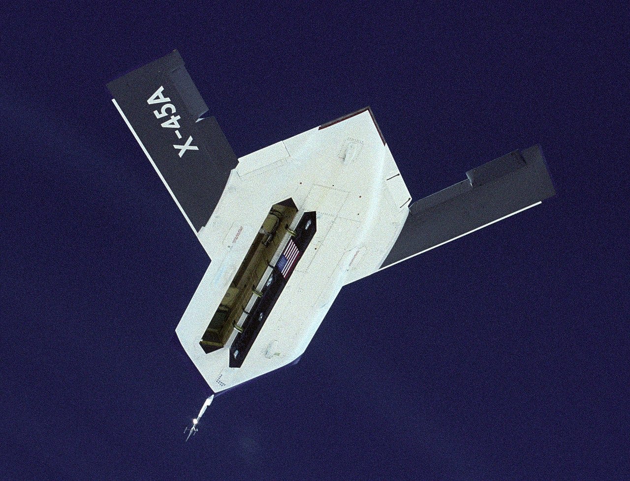 A sharply angular drone in flight against a grainy blue sky, with bomb bay open. A sharply angular drone in flight against a grainy blue sky, with bomb bay open.