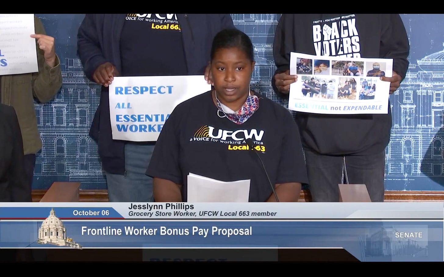 A black woman in a black tee shirt with white text reading UFCW Local 663 stands behind a podium, speaking A black woman in a black tee shirt with white text reading UFCW Local 663 stands behind a podium, speaking