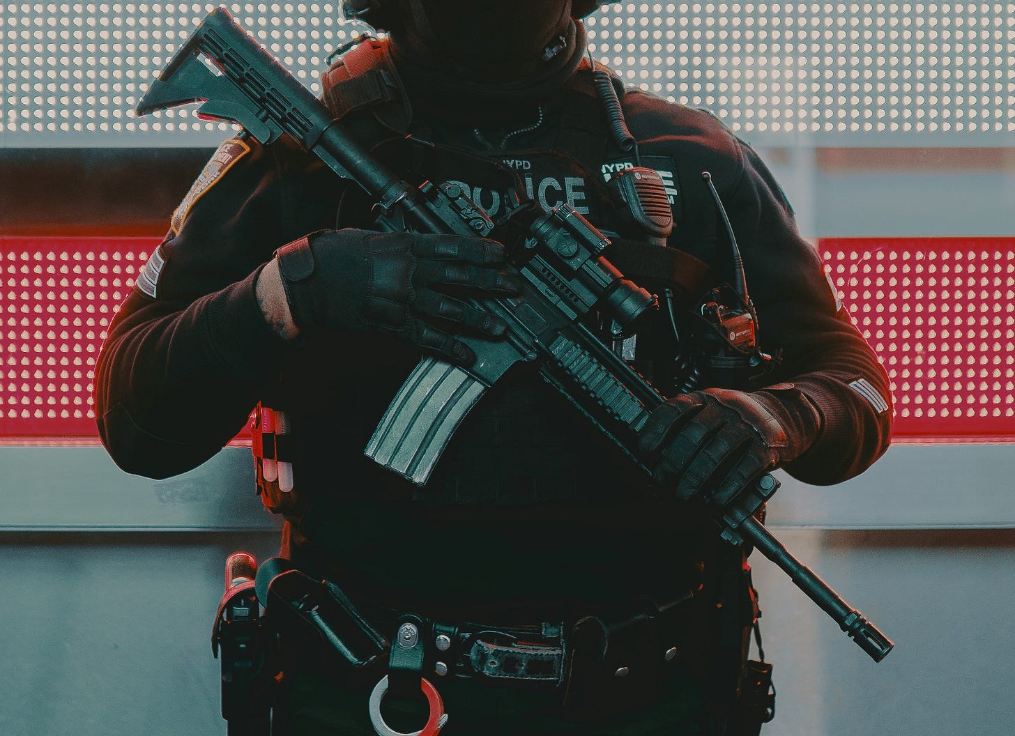 Cropped photo of a heavily armed police officer in front of a neon red and white background.