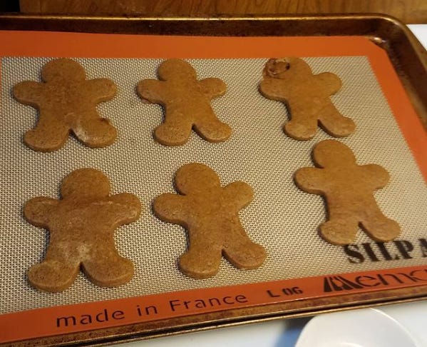 Six gingerbread cookies shaped like people laying on a silicone baking mat. The cookie on the upper right looks like its head exploded in the oven. Six gingerbread cookies shaped like people laying on a silicone baking mat. The cookie on the upper right looks like its head exploded in the oven.