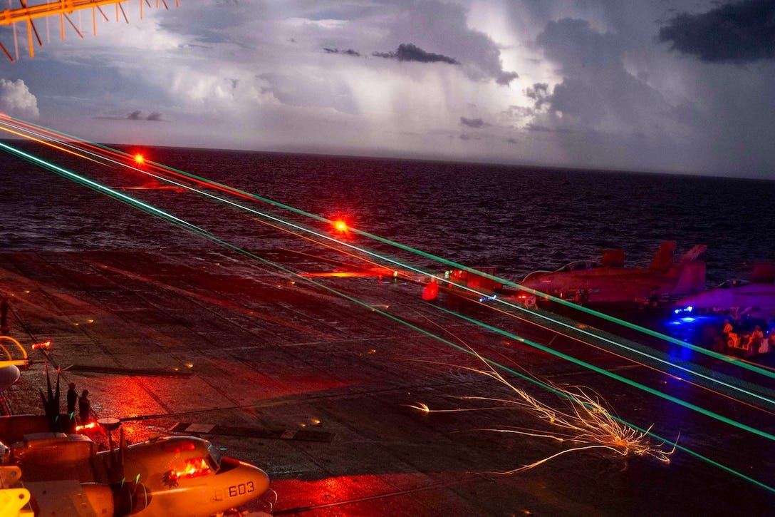 A Navy F/A-18E Super Hornet lands on the flight deck of the USS Carl Vinson in the South China Sea, Oct. 5, 2021. An aircraft lands on the flight deck of a ship at sea illuminated by colorful lights.