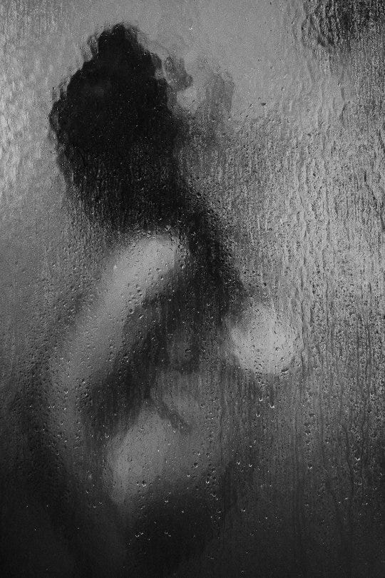 Descrição da imagem: em preto e branco. O corpo de uma mulher de perfil num box de chuveiro, onde ela está de lado, olhando pra cima. Cabelos negros e uma fonte de luz vinda de cima, mais para a direita da foto. Descrição da imagem: em preto e branco. O corpo de uma mulher de perfil num box de chuveiro, onde ela está de lado, olhando pra cima. Cabelos negros e uma fonte de luz vinda de cima, mais para a direita da foto.